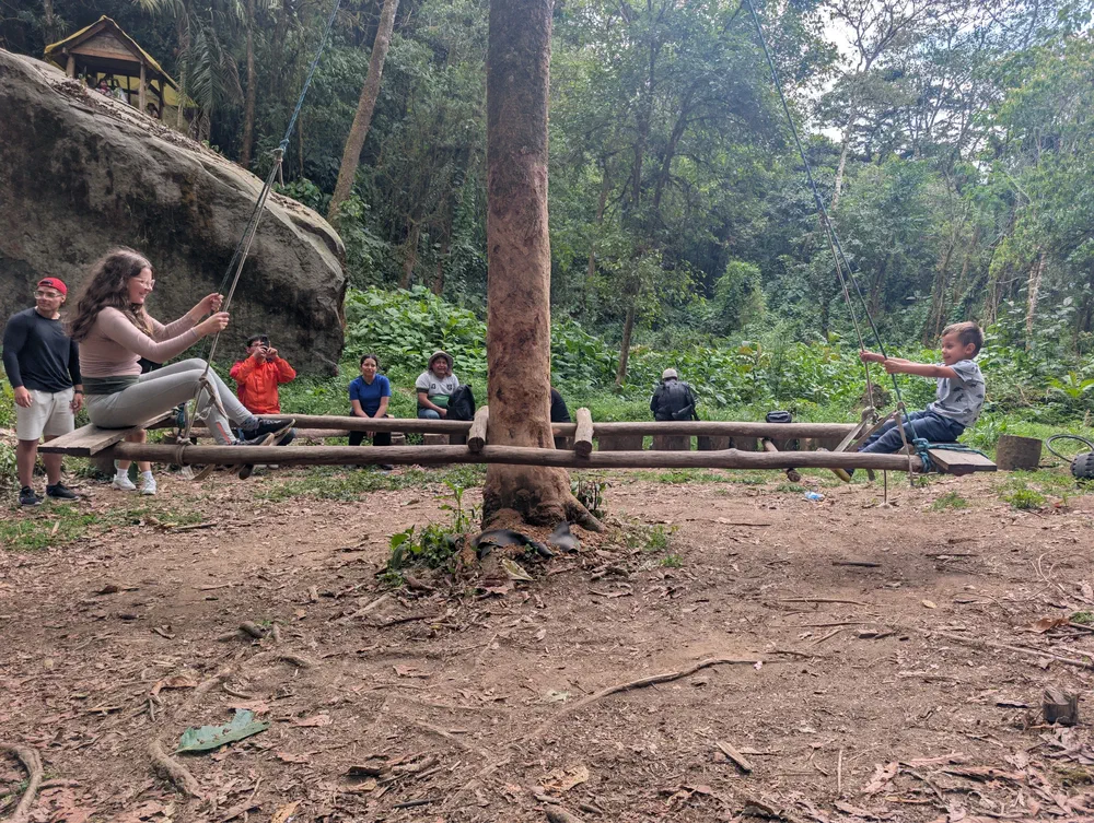 Two children on either end of a long log seesaw suspended by ropes from a tree, with other visitors watching from a bench in the clearing