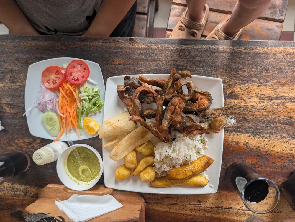 An overhead view of a plate of fried trout with rice, yuca, fried potatoes and sweet plantain, beside a side salad and a bowl of green llajwa