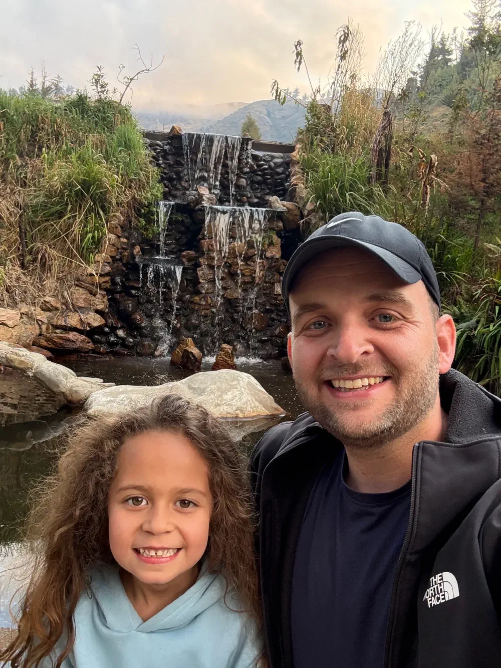 A man and a young girl smiling for a selfie in front of a landscaped stone waterfall spilling into a pool, with hills and low cloud behind them
