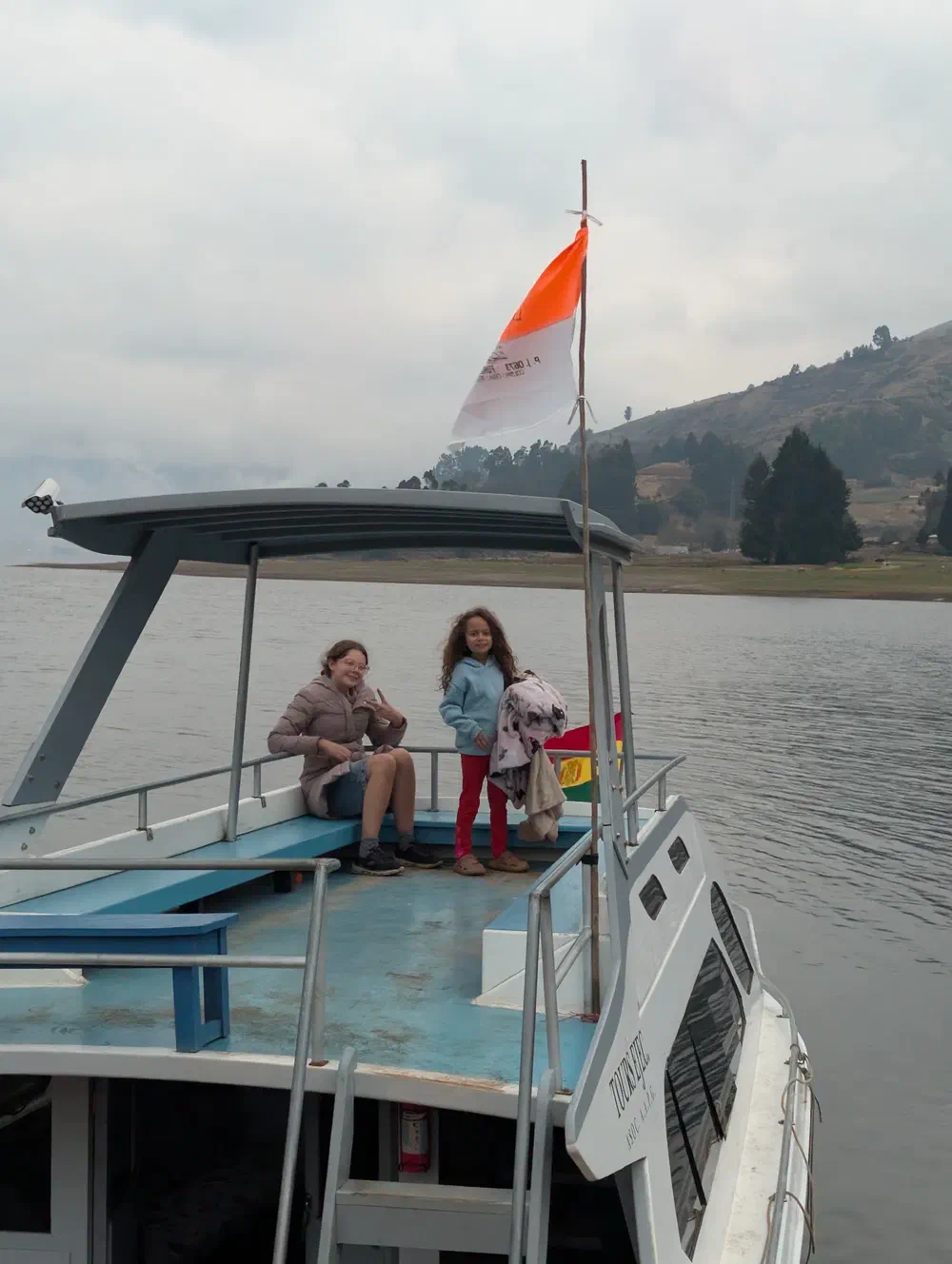 My daughters standing on the open upper deck of a small tour boat on Corani lake, with a Bolivian flag on the rail and pine-covered hills across the grey water