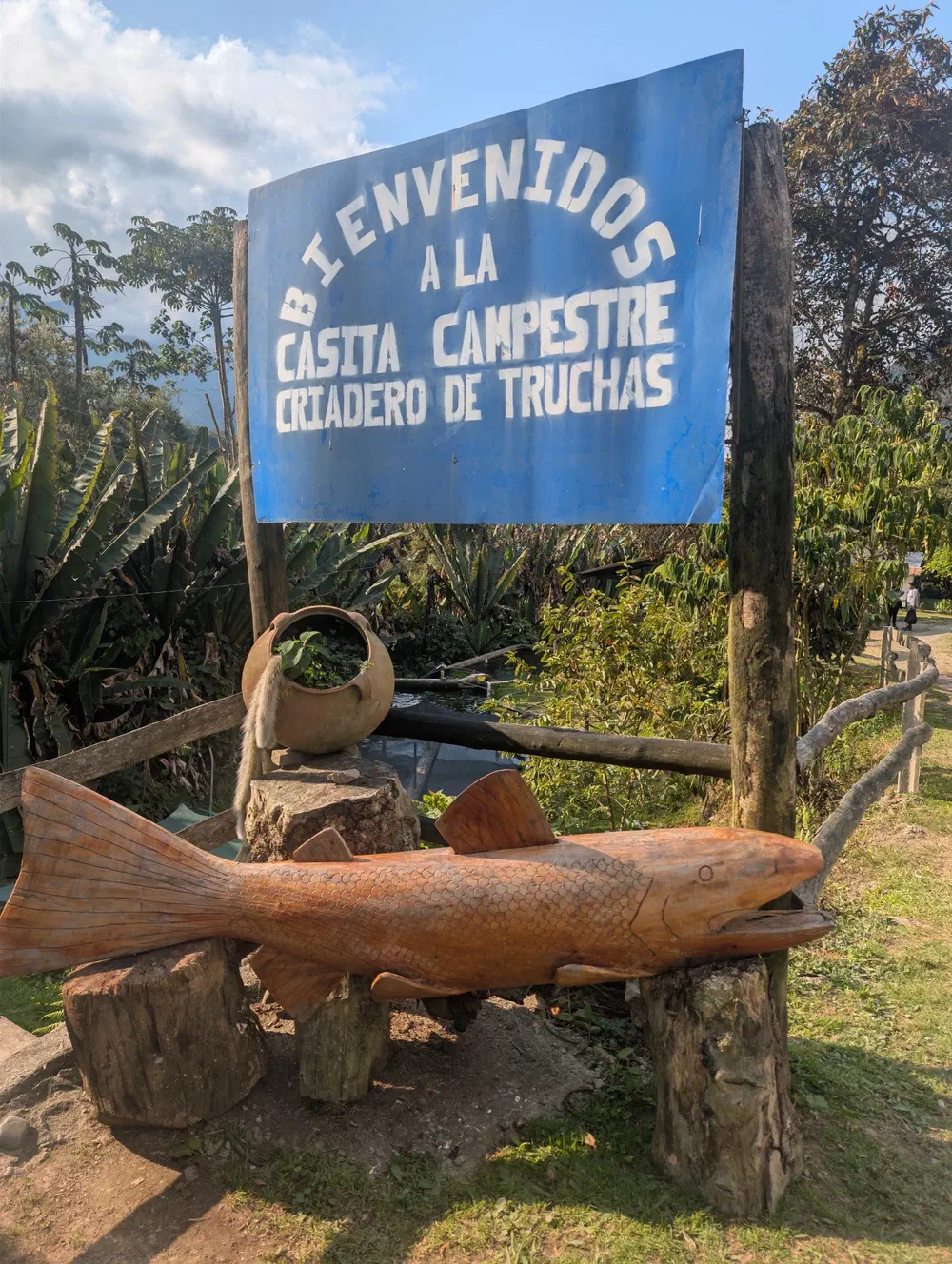 A hand-painted blue sign reading Bienvenidos a la Casita Campestre Criadero de Truchas, mounted above a large carved wooden trout on log legs