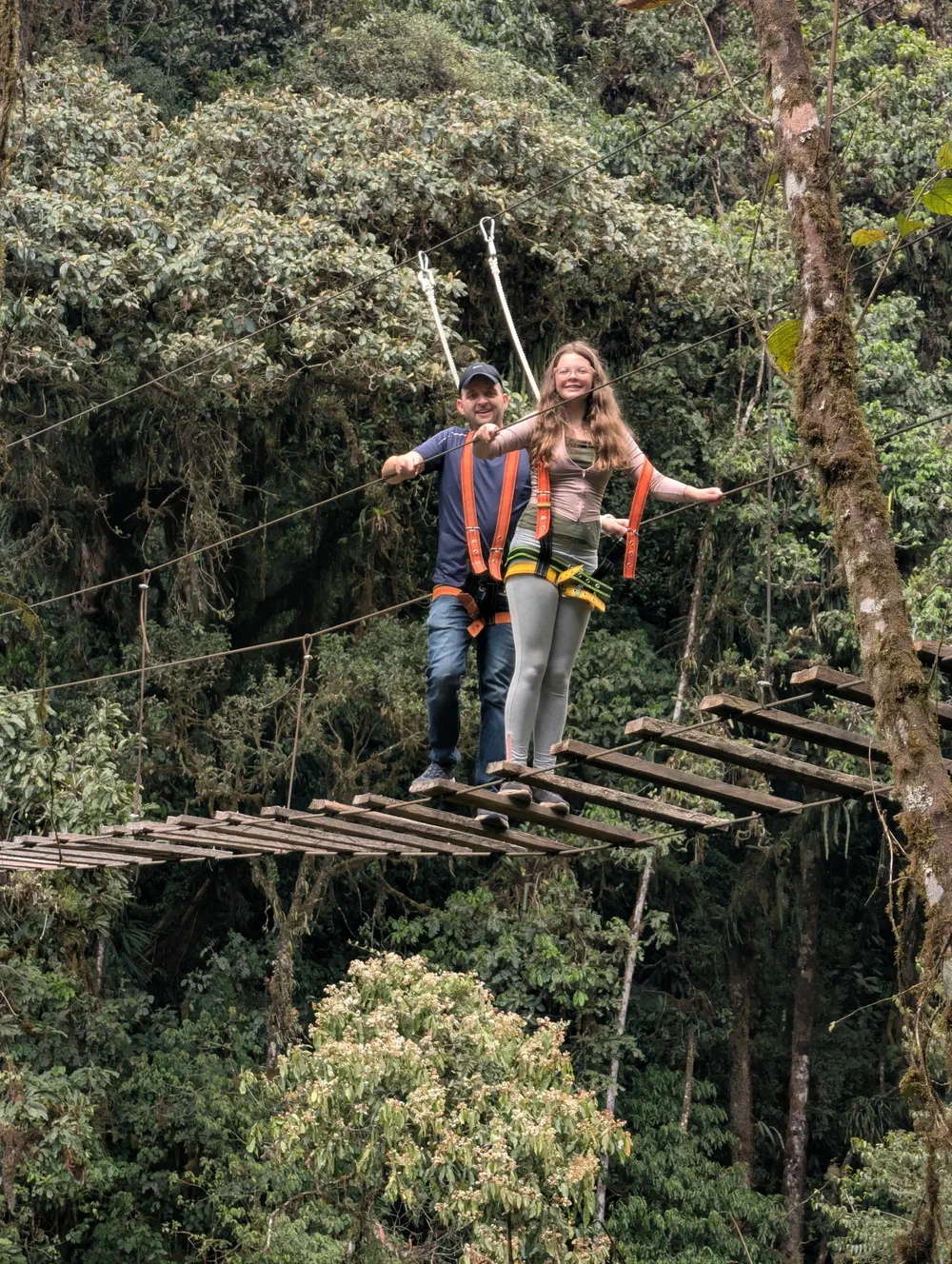 My daughter and I climbing harnesses standing on a long, narrow suspension bridge of spaced wooden slats, strung high through the cloud forest canopy
