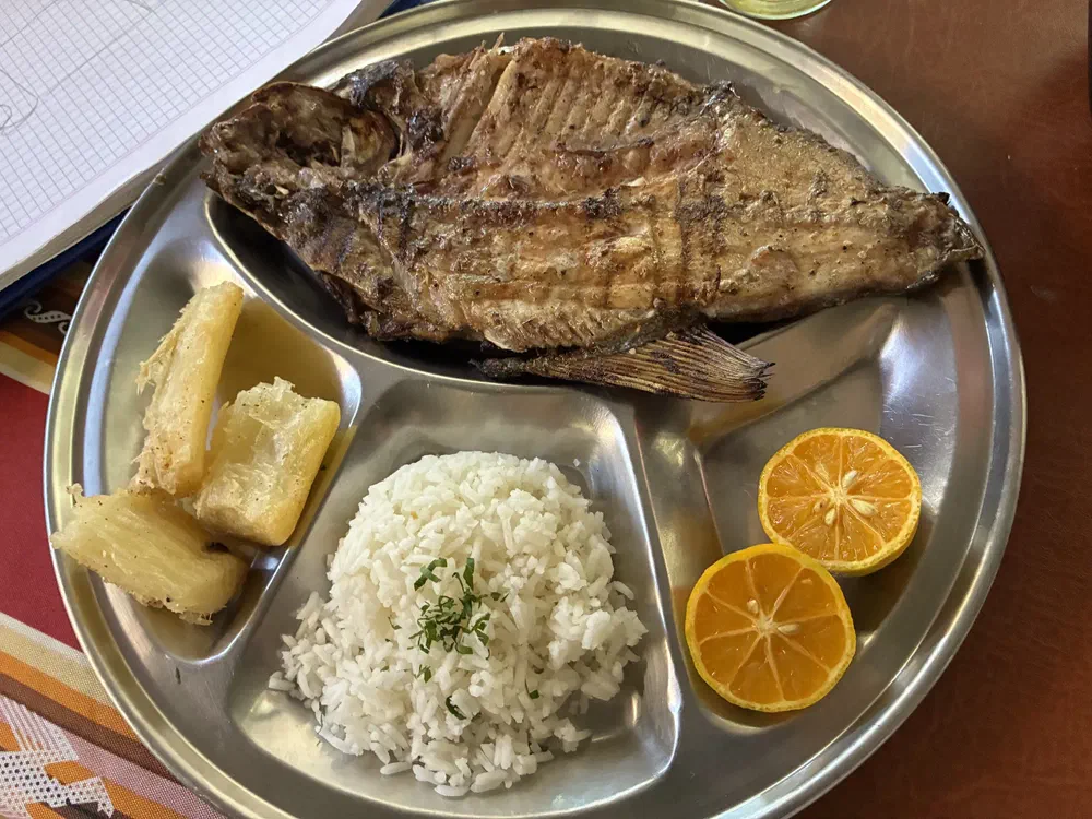 A whole grilled fish on a divided steel tray with white rice, yuca and halved limes