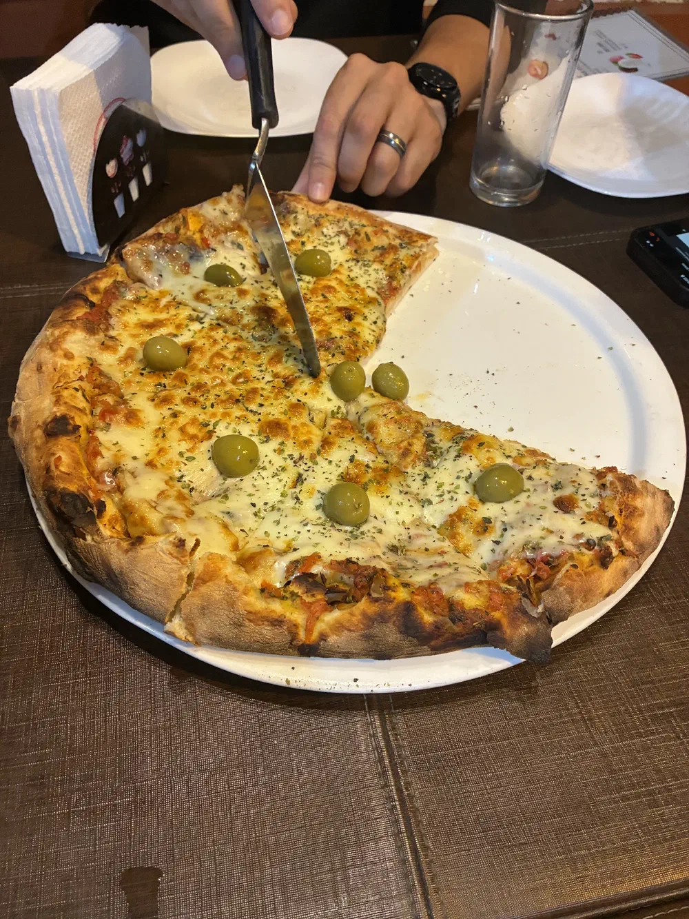 A mozzarella pizza topped with green olives and oregano, part-sliced, with a hand drawing a knife through it